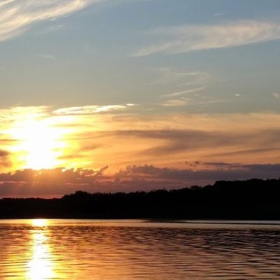 Sunset over water with reflections and a silhouette of trees on the horizon.