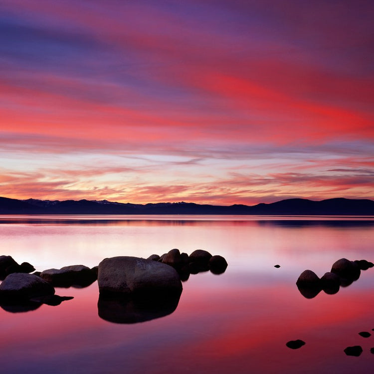 Sunset over a tranquil lake with vibrant sky reflection and rocks in water.