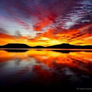 Sunset with vivid orange clouds reflected in calm lake with silhouetted hills.