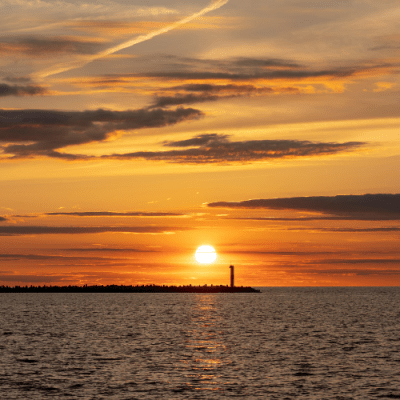 Sunset over water with clouds and distant silhouette of a lighthouse.