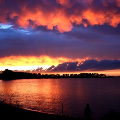 Person silhouetted against vibrant sunset over water with dramatic clouds.