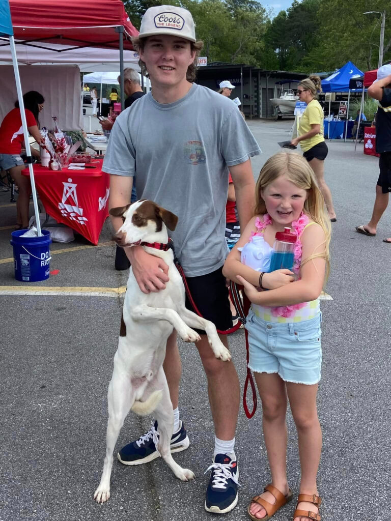 Smiling boy with a dog and a girl holding a drink at an outdoor event with colorful tents and people in the background.