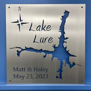 Metal sign with Lake Lure map, compass rose, and "Matt & Haley, May 23, 2023" on brushed metal, mounted on a blue wall.