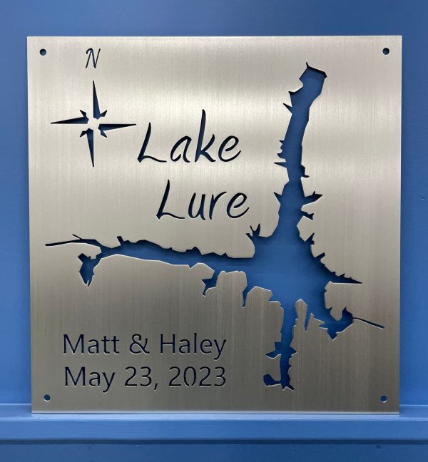 Metal sign with Lake Lure map, compass rose, and "Matt & Haley, May 23, 2023" on brushed metal, mounted on a blue wall.