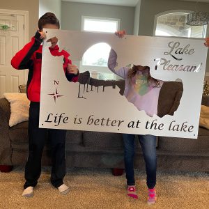 Children holding a Lake Pleasant metal sign with "Life is better at the lake" text in a cozy living room.