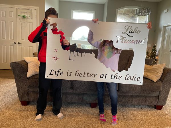 Children holding a Lake Pleasant metal sign with "Life is better at the lake" text in a cozy living room.