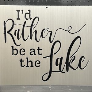 "I'd rather be at the lake" text on a brushed metal sign with decorative script against a perforated backdrop.