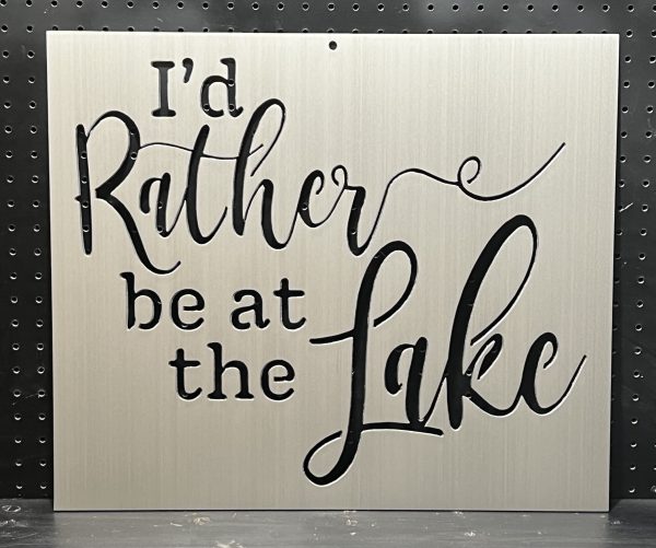 "I'd rather be at the lake" text on a brushed metal sign with decorative script against a perforated backdrop.