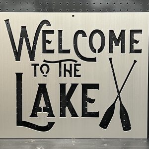 "Welcome to the Lake" metal sign with crossed oars cutout, set against a perforated backdrop.