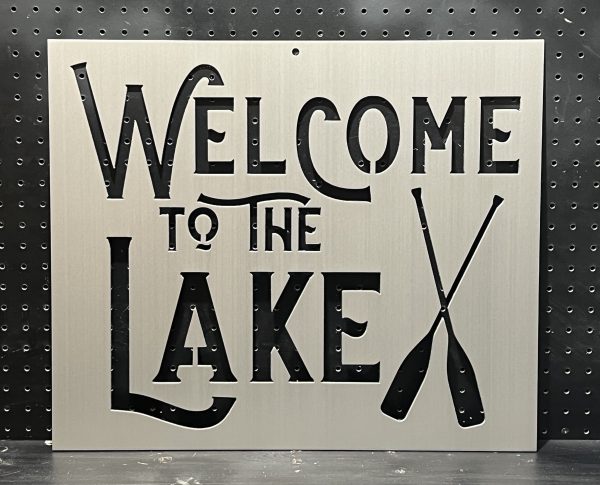 "Welcome to the Lake" metal sign with crossed oars cutout, set against a perforated backdrop.