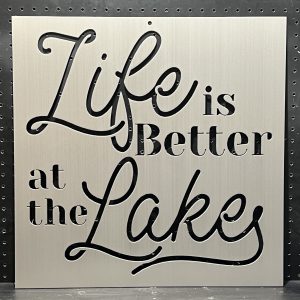 "Life is Better at the Lake" text cutout on brushed metal sign with decorative script against a perforated background.