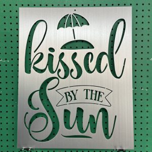 Metal sign with "kissed by the sun" cutout design and umbrella motif on green pegboard background.