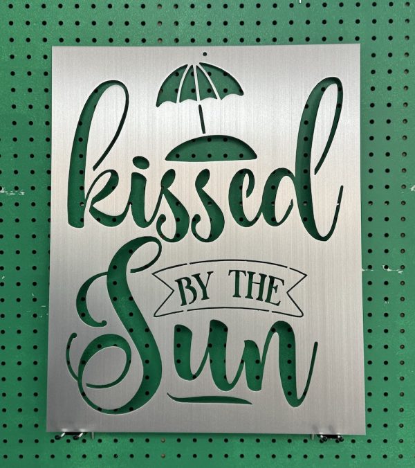 Metal sign with "kissed by the sun" cutout design and umbrella motif on green pegboard background.