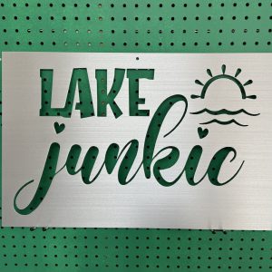 "Lake Junkie" metal sign with sun and wave cutout, set against a green perforated background.