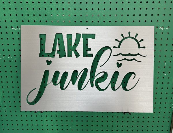 "Lake Junkie" metal sign with sun and wave cutout, set against a green perforated background.