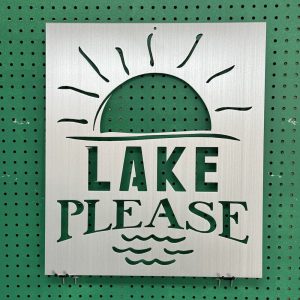 Metal sign with "Lake Please" text and sun design, mounted on a green pegboard.