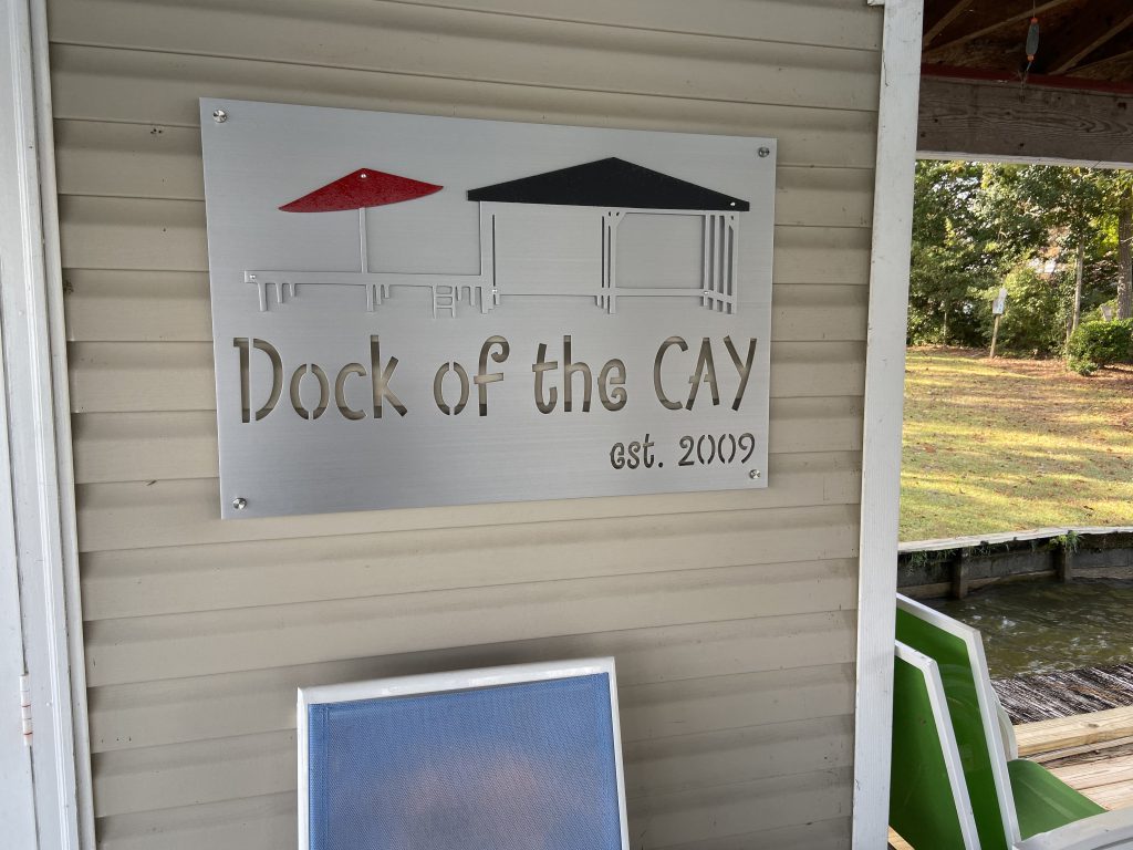 Custom metal sign reading "Dock of the CAY, est. 2009" with red and black umbrellas on a beige wall.