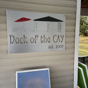 Custom metal sign reading "Dock of the CAY, est. 2009" with red and black umbrellas on a beige wall.