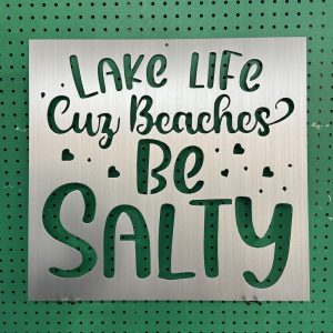 Metal sign with "Lake Life Cuz Beaches Be Salty" text and heart accents on a green pegboard background.