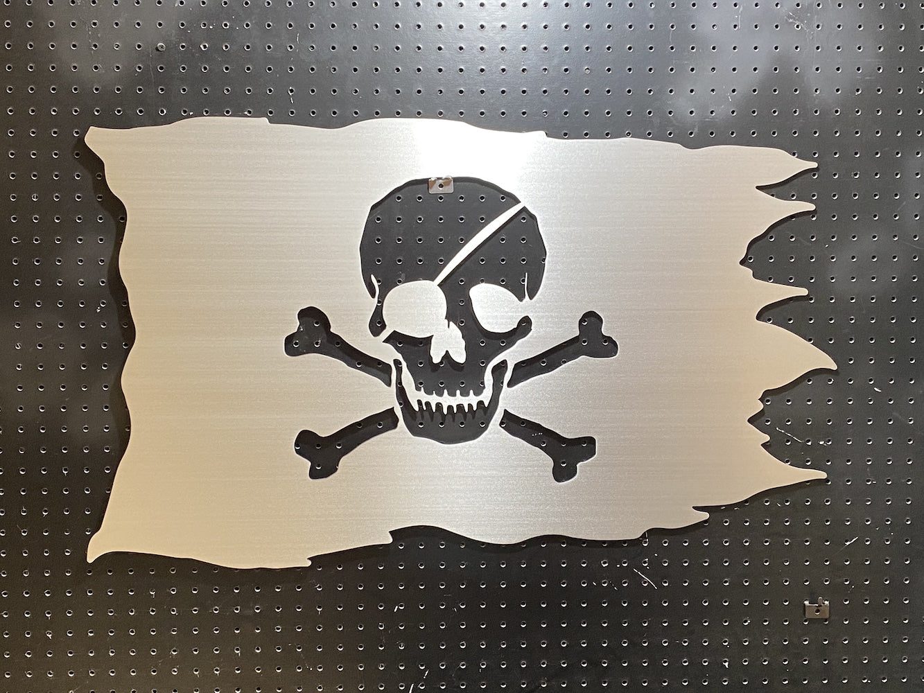 Metal pirate flag design with a skull and crossbones cutout, mounted on a black pegboard.