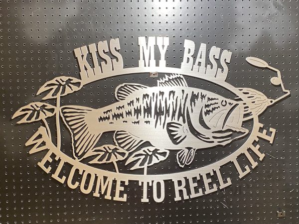 "Kiss My Bass" metal sign with a fish design, featuring "Welcome to Reel Life" text on dark perforated background.