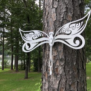 White metal dragonfly art with ornate wings on a tree, set against a lush forest and serene lake backdrop.