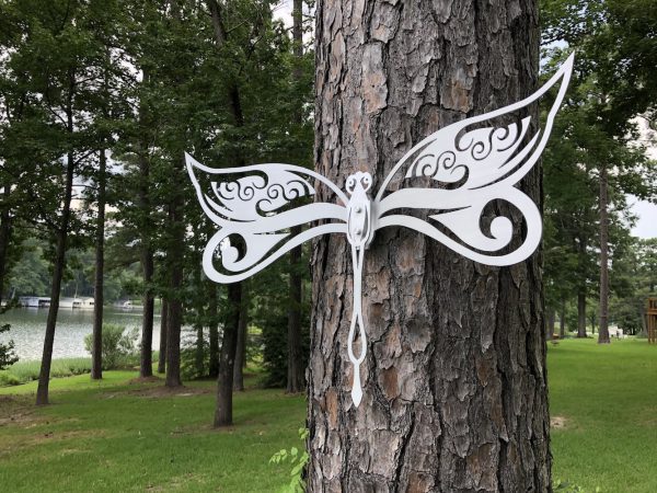 White metal dragonfly art with ornate wings on a tree, set against a lush forest and serene lake backdrop.