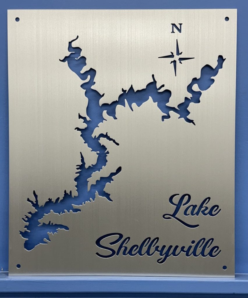 Lake Shelbyville cutout metal map with compass rose on brushed silver, mounted on a blue wall.