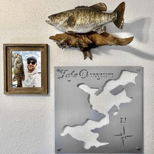 Lake Oneegum metal map with compass rose, mounted beside a fish mount and framed fishing photo on a light wall.