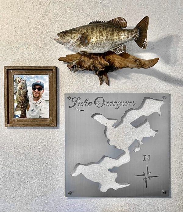 Lake Oneegum metal map with compass rose, mounted beside a fish mount and framed fishing photo on a light wall.