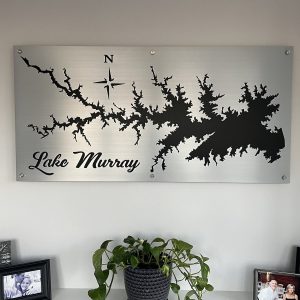 Lake Murray metal map with compass rose, displayed above a shelf with family photos and a potted plant.