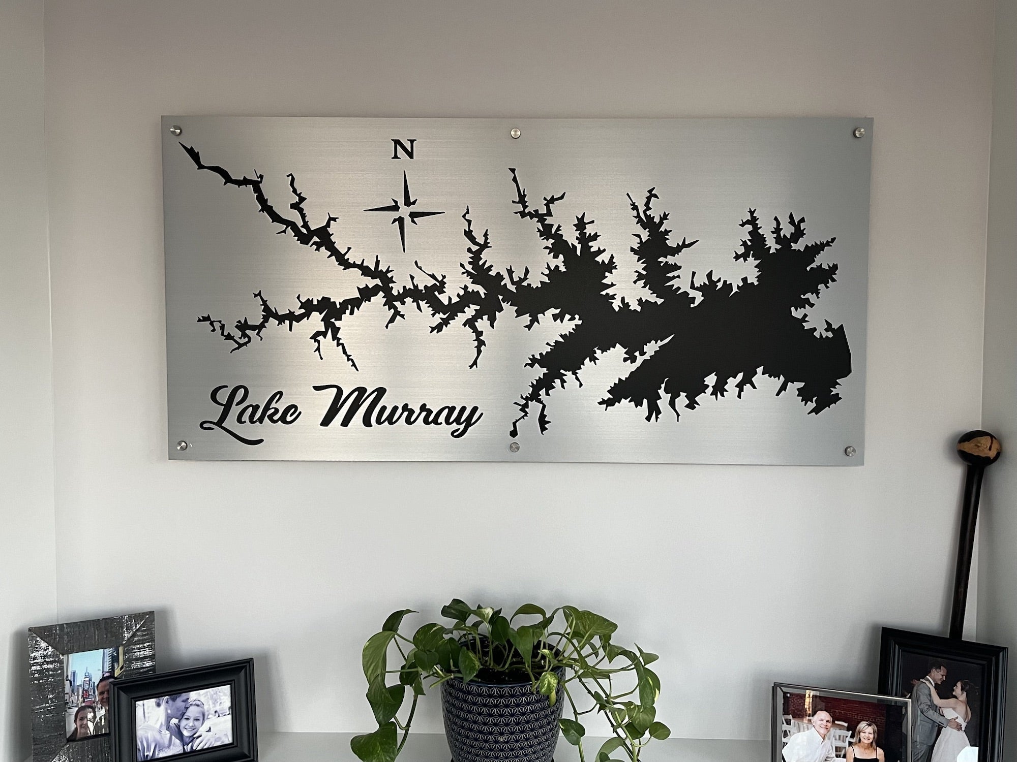 Lake Murray metal map with compass rose, displayed above a shelf with family photos and a potted plant.