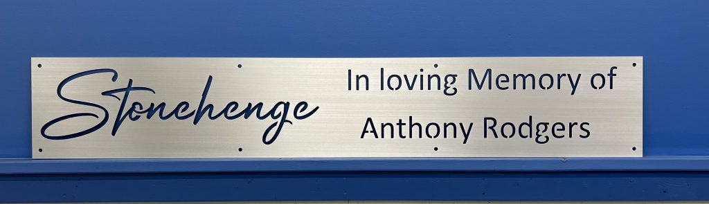 Brushed metal sign on blue wall, reads "Stonehenge In loving Memory of Anthony Rodgers."