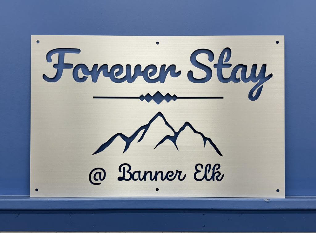 Metal sign with "Forever Stay @ Banner Elk" and mountain design on a blue wall.