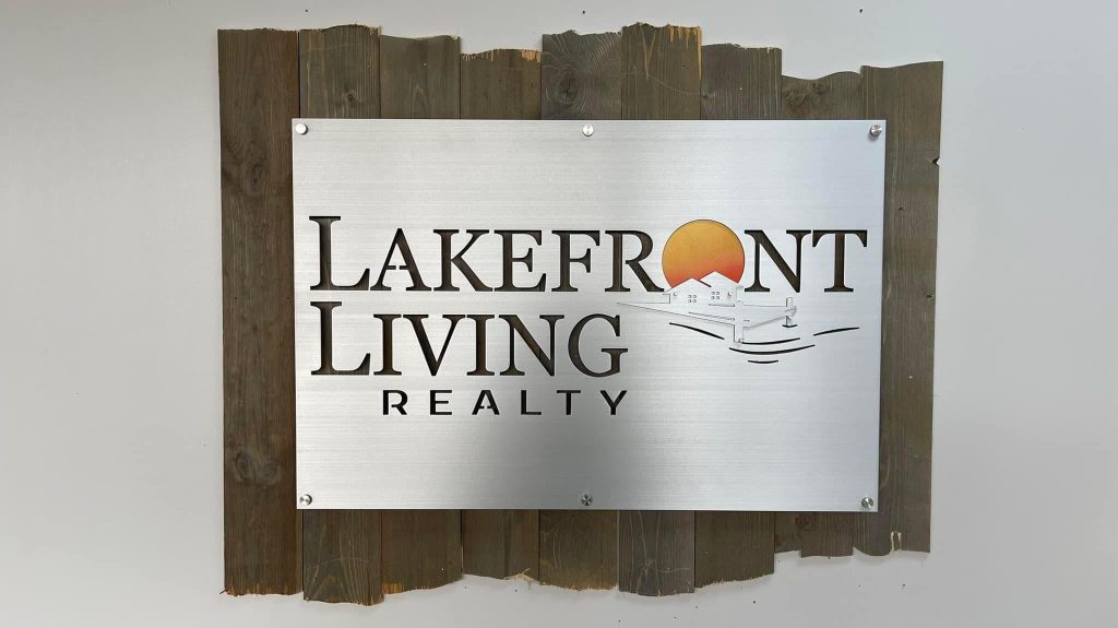 Lakefront Living Realty metal sign with house and sunset design on rustic wood backdrop.