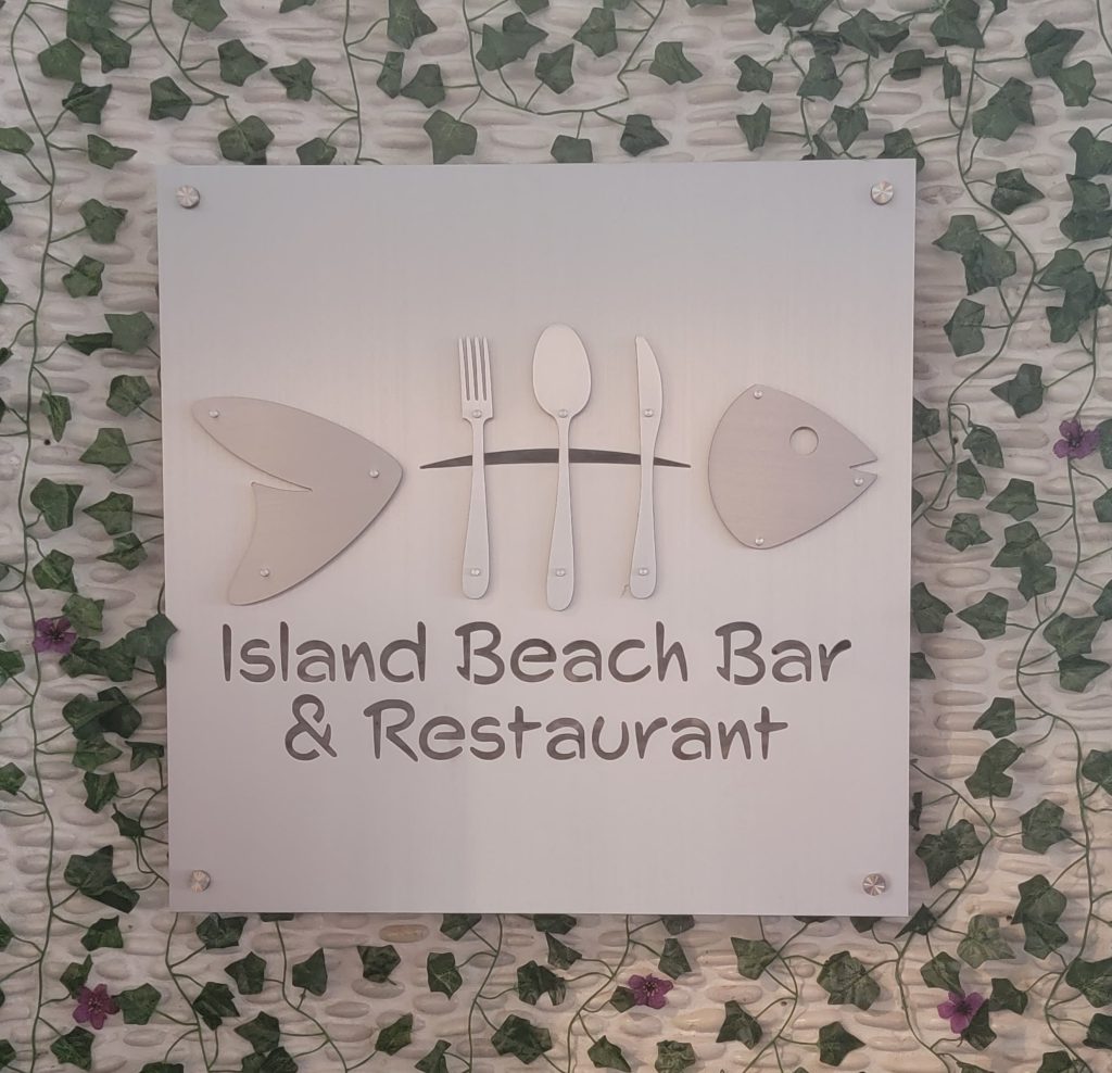 Island Beach Bar & Restaurant sign with knife and fork fish design, on a leafy green wall.