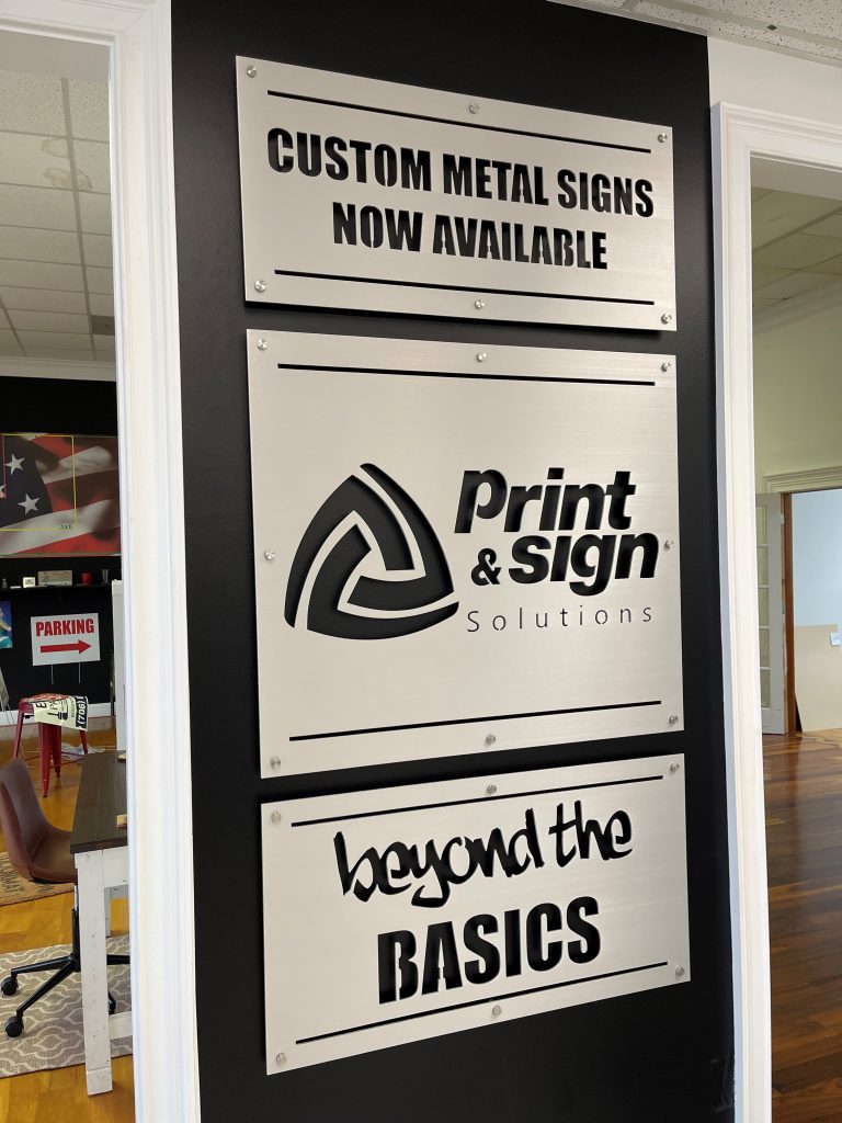 Custom metal signs displayed on a black wall: "Custom Metal Signs Now Available", "Print & Sign Solutions", "Beyond the Basics".