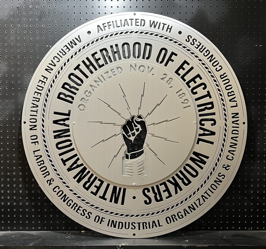 Circular metal sign for International Brotherhood of Electrical Workers, organized Nov. 28, 1891, on a pegboard background.