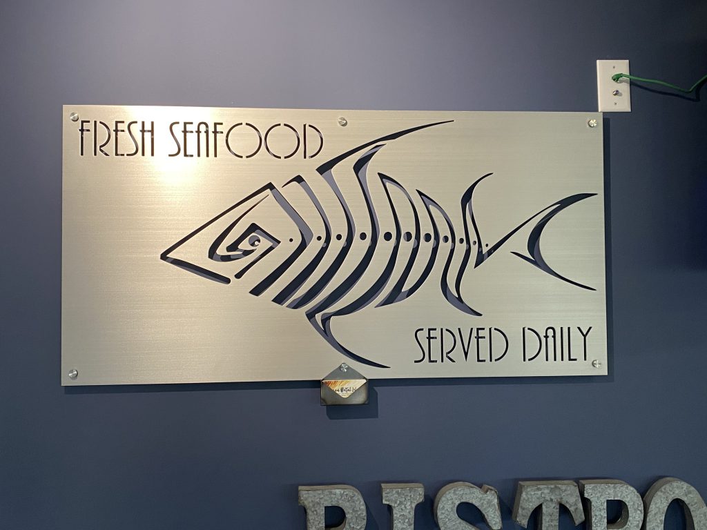 Metal sign with stylized fish design and text "Fresh Seafood Served Daily" on a blue wall.