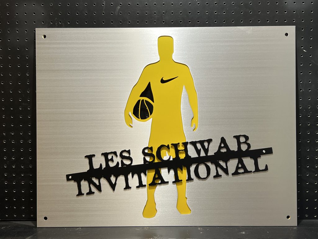 Les Schwab Invitational metal sign with basketball player silhouette and Nike logo, mounted on a pegboard.