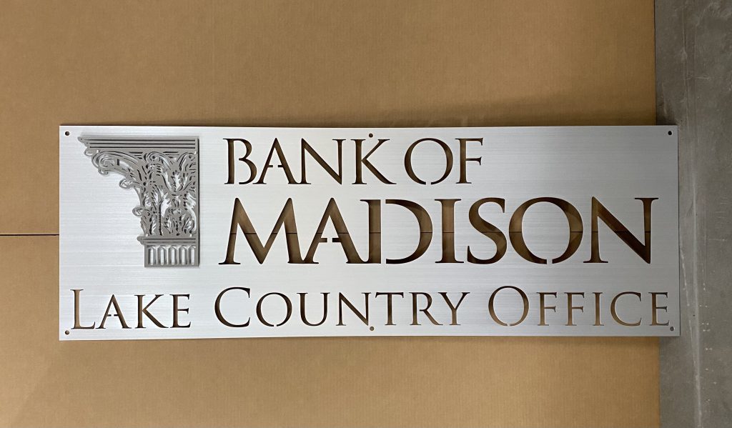 Bank of Madison Lake Country Office metal sign with ornate column detail, mounted on a beige wall.