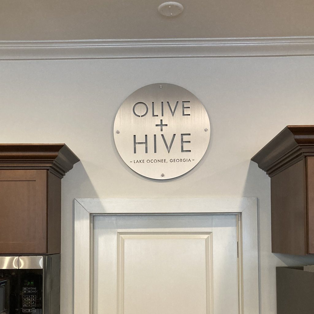 Round metal sign reading "Olive + Hive, Lake Oconee, Georgia" above a doorway with wooden cabinets on either side.