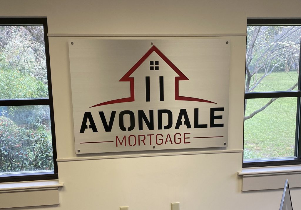 Avondale Mortgage sign with house logo, mounted between two windows overlooking greenery.