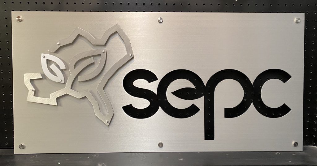 Brushed metal sign with "sepc" logo and abstract design on a perforated black background.