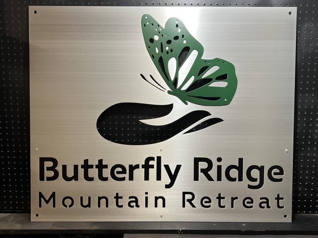 Butterfly Ridge Mountain Retreat metal sign with butterfly design on brushed silver, mounted on a black pegboard.