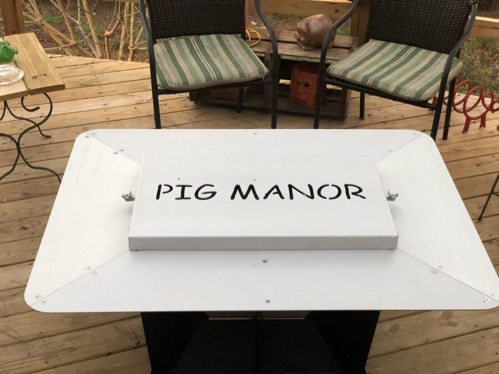 Customizable metal fire pit cover with "Pig Manor" text on a wooden porch setting.