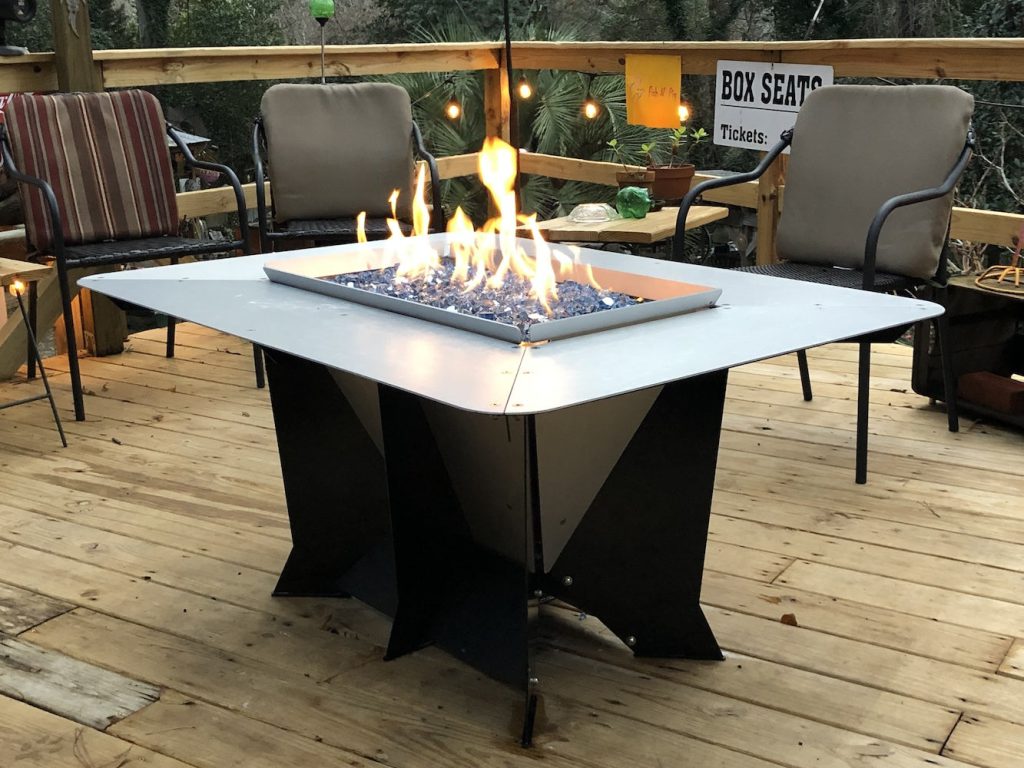 Modern gas fire pit with blue stones on a wooden deck, surrounded by chairs and a "Box Seats" sign in the background.