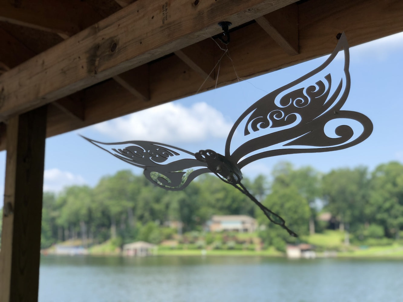 Metal dragonfly with intricate design hanging under a wooden dock, overlooking a serene lake and tree-lined shore.