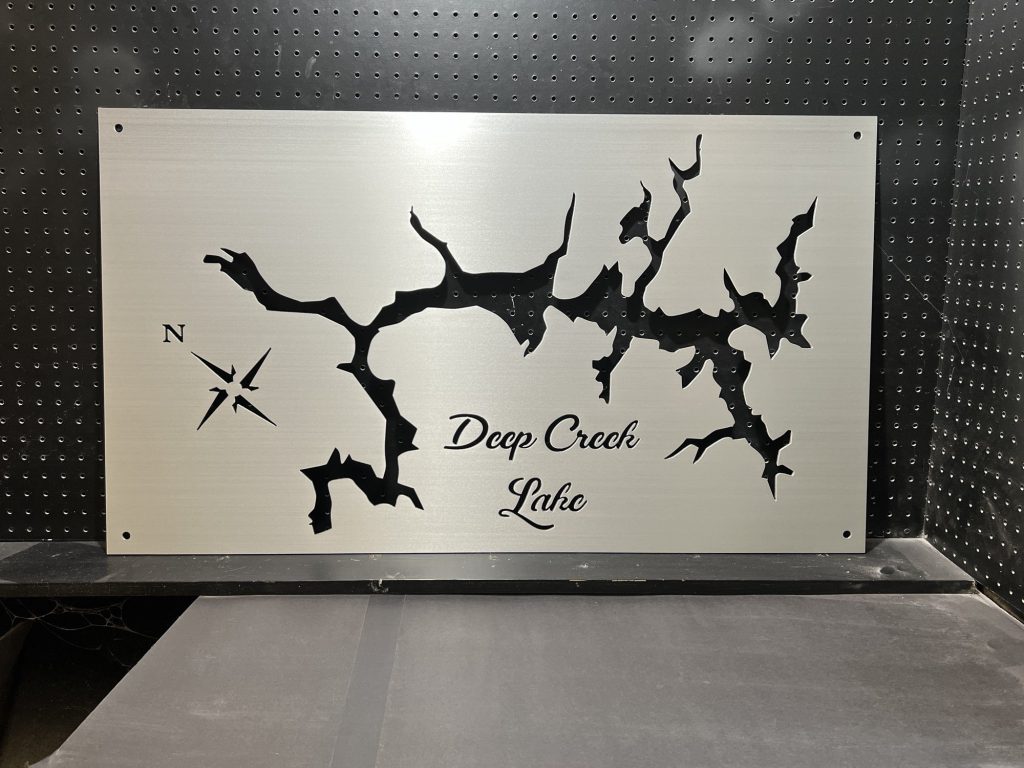 Metal map of Deep Creek Lake with compass rose, mounted on a perforated black background.