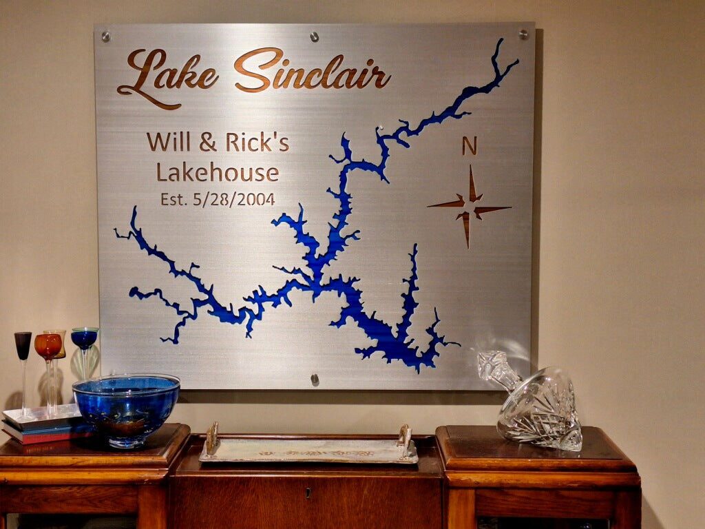 wp-image47163052851494.png Lake Sinclair metal map with "Will & Rick's Lakehouse Est. 5/28/2004" and compass rose, displayed on a wooden side table.